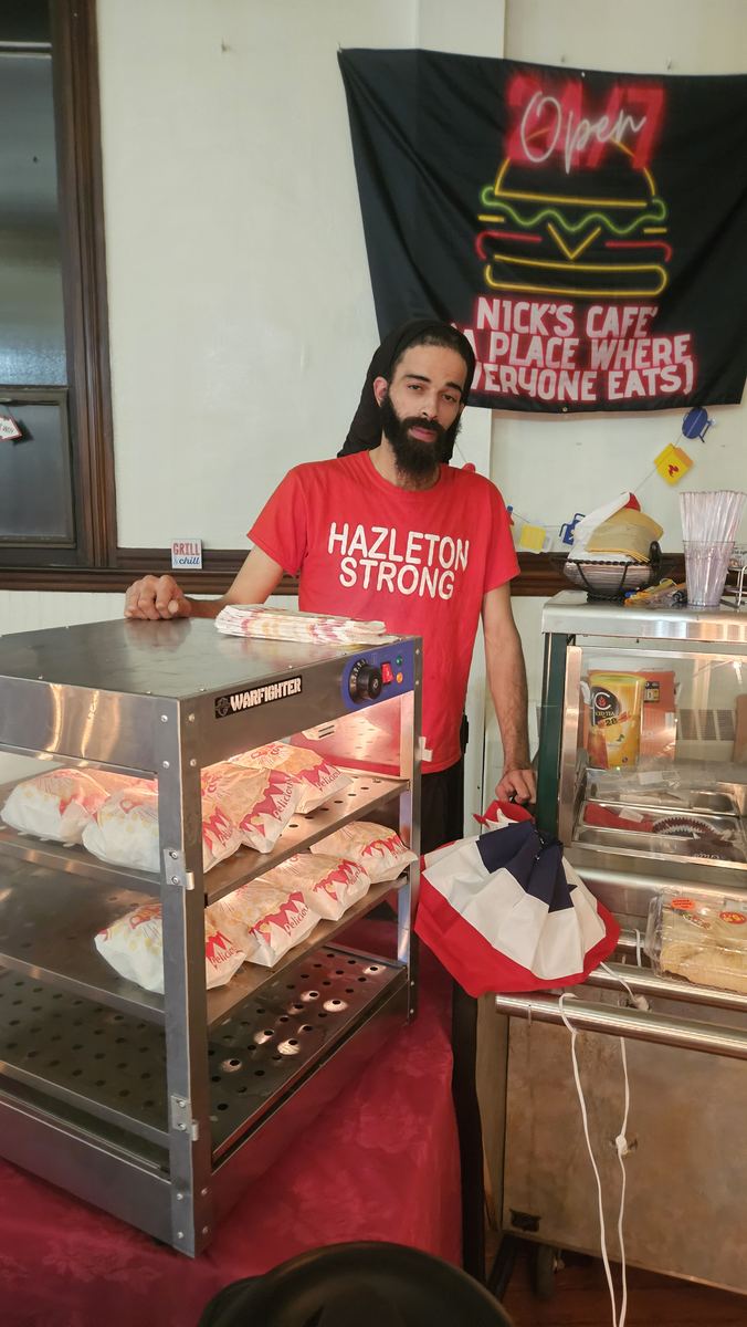 Chef Nick behind the Nick's Café food warmer, under a banner reading Nick's Café, a place where everyone eats